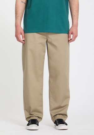 Personne portant un pantalon beige ample, des baskets noir et blanc, et un t-shirt bleu sarcelle, debout devant un fond blanc uni.