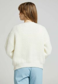 Pull crème flou avec des épaules tombantes et un ourlet côtelé. Porté avec un pantalon bleu clair, mettant en avant une texture douce et une coupe décontractée.