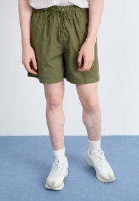 Osoba v olivově zelených šortkách na stažení, bílých sportovních botách a bílých ponožkách stojí na modré podlaze na jednotném pozadí.