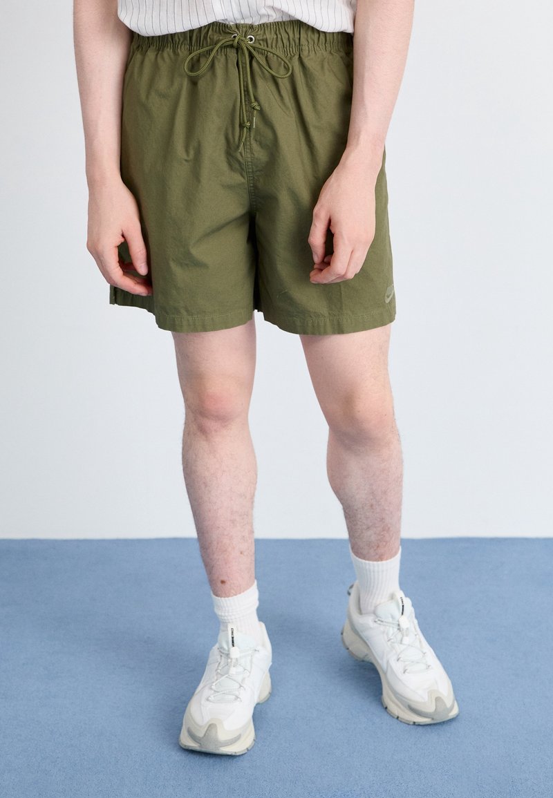 Osoba v olivově zelených šortkách na stažení, bílých sportovních botách a bílých ponožkách stojí na modré podlaze na jednotném pozadí.