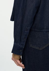 Κοντινό πλάνο ενός χεριού που φοράει σκούρο μπλε τζιν πουκάμισο με κουμπωμένο μανίκι πάνω από μακρυμάνικη μπλούζα σε ταμπά χρώμα, δίπλα στην τσέπη σκουρόχρωμου μπλε τζιν παντελονιού.