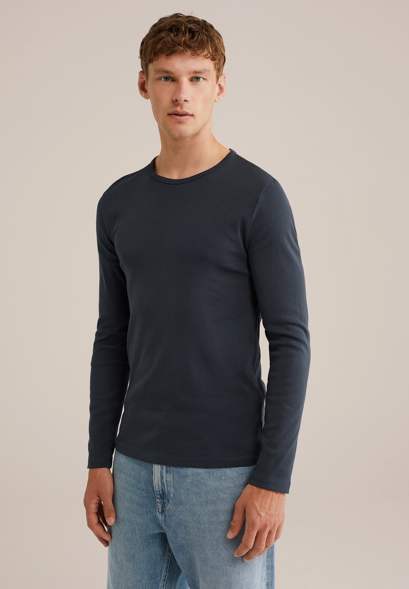 Vyras, apsirengęs prigludusia tamsiai mėlyna ilgomis rankovėmis marškiniais ir šviesiai mėlynais džinsais, stovi prieš neutralų foną.