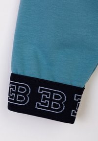 Tessuto di cotone teal con polsino a coste nero. Il polsino presenta un logo ricamato in bianco "EB". Texture liscia, design aderente.