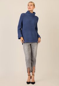 Blauer gerippter übergroßer Pullover mit umgeschlagenem Kragen, kombiniert mit grauen, verwaschenen Jeans mit Fransen und Verzierungen, getragen mit schwarzen Absatzschuhen.