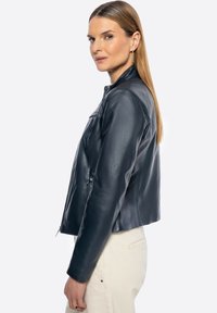 Marineblaue Lederjacke, taillierte Passform, hoher Kragen, mit Reißverschlüssen versehene Taschen und glatte Textur, kombiniert mit hellen, neutralen Hosen.