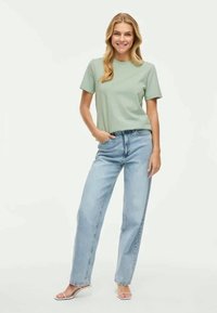 Gaiši zaļš kokvilnas T-krekls ar īsām piedurknēm, kombinēts ar augstās jostas klasiski gaiši ziliem džinsiem un baltām zābaku sandālēm.