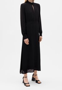 Schwarzes, langärmeliges Kleid mit hohem Ausschnitt und Schlitz vorne. Hat eine taillierte Taille und einen fließenden Saum. Getragen mit schwarzen Blockabsatzsandalen.