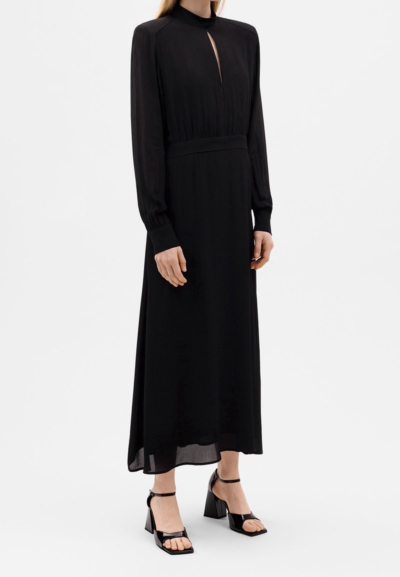 Schwarzes, langärmeliges Kleid mit hohem Ausschnitt und Schlitz vorne. Hat eine taillierte Taille und einen fließenden Saum. Getragen mit schwarzen Blockabsatzsandalen.