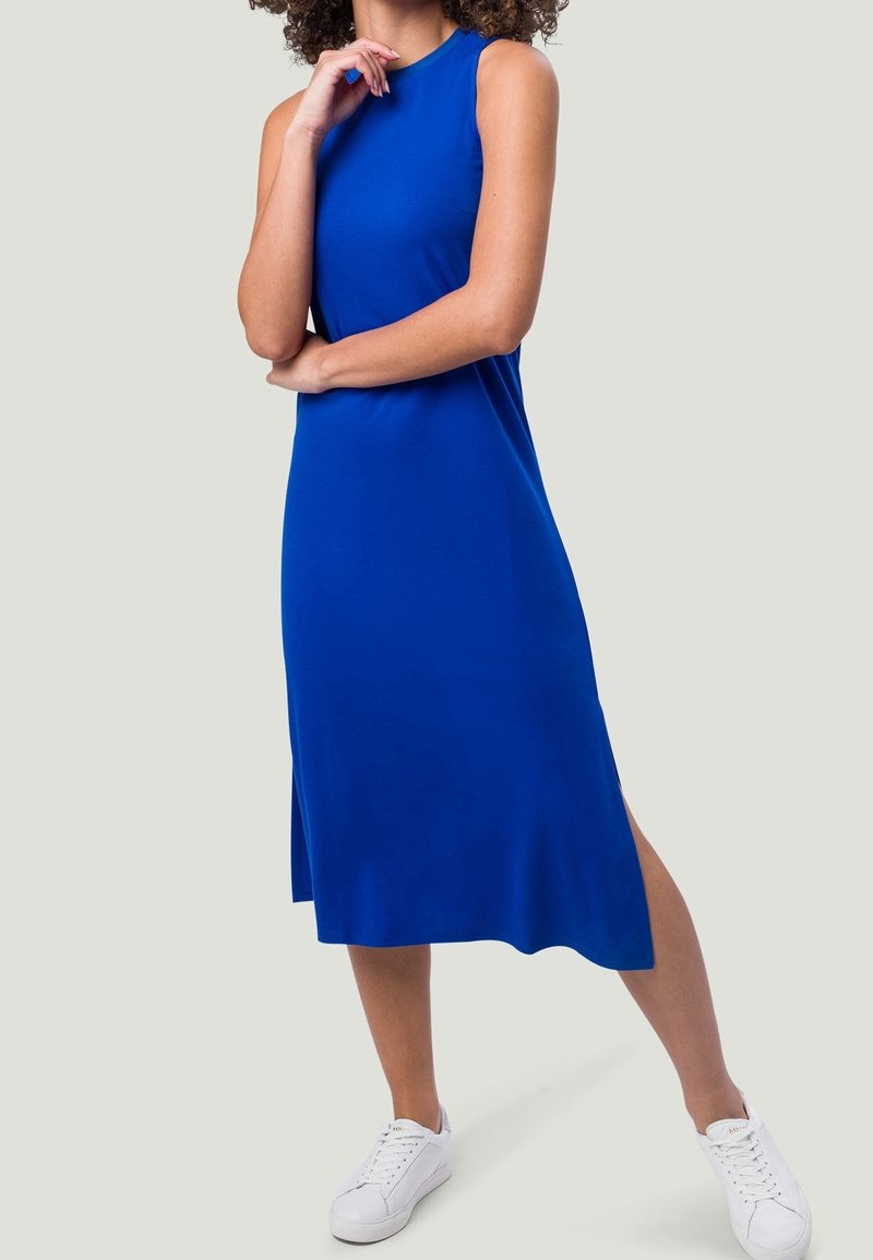Femme portant une robe bleu cobalt sans manches, arrivant au genou, associée à des baskets blanches, debout avec une jambe légèrement avancée.