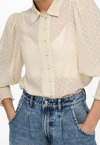 Przeźroczysta, jasnobeżowa bluzka w kropki z zapięciem na guziki z przodu i bufiastymi rękawami, w zestawieniu z niebieskimi dżinsami z wysokim stanem.