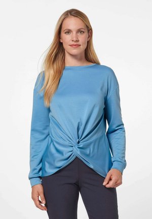 Lichtblauw langwerpig shirt met een knoopdetail bij de taille, voorzien van een ronde halslijn en zachte stof, gecombineerd met donkere broek.