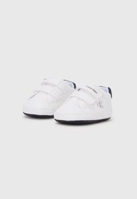 Λευκά υφασμάτινα παιδικά παπούτσια με λουράκια βέλκρο και μαύρη λαστιχένια σόλα. Διαθέτουν λογότυπο Calvin Klein σε ασημί στο πλάι.