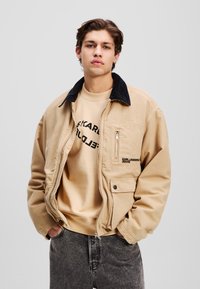 Beige bomullsjakke med svart kordfløyelskrage, glidelås foran, lommepreg, og brodert logo. Båret over en beige genser og mørke jeans.