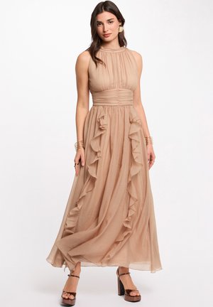 Mujer con vestido largo beige sin mangas y de cuello alto, con detalles de volantes y tacones de plataforma, posando frente a un fondo blanco liso.