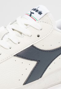 Vit lädersneaker med mörk marinblå accenter. Har texturerad yta, perforeringar och platta vita skosnören. Märke på tungan.