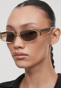 Ochelari de soare bej transparenți cu rame geometrice și lentile închise la culoare, având un design elegant și accente hardware minime.