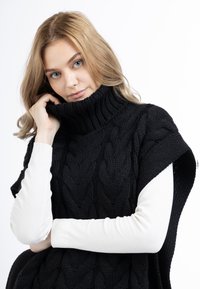Schwarzer Strickpullover mit hohem Rollkragen, ausgestattet mit einem Zopfmuster und offenen Seiten, über einem weißen Langarmshirt getragen.