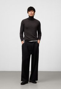 Donkergrijze coltrui, lichtgewicht breiwerk, gecombineerd met zwarte wijde broek. Model staat zelfverzekerd met de handen in de zakken, in zwarte schoenen.