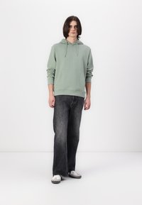 Felpa verde chiaro con tasca frontale, abbinata a jeans svasati grigio scuro. La persona indossa sneaker bianche e grigie. Sfondo semplice.