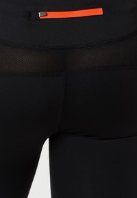 Leggings deportivos negros con un panel de malla texturada y un bolsillo con cremallera naranja en la parte posterior. Hechos de un material elástico y ajustado.