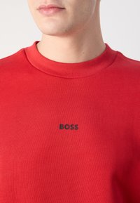 Gros plan sur une personne portant un sweat-shirt rouge à col rond avec le mot "BOSS" imprimé en noir sur la poitrine.