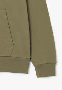 Gros plan sur la manche et la poche avant d'un sweat-shirt vert olive avec poignets et ourlet côtelés sur fond uni.