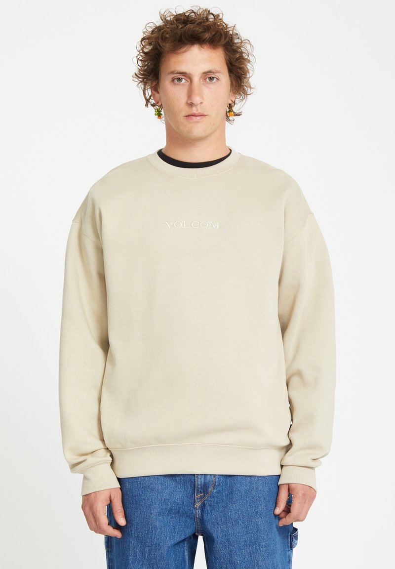 Beige crewneck sweatshirt with ribbed cuffs and hem, featuring embroidered "VOLCOM" logo on chest. Smooth fabric and relaxed fit.