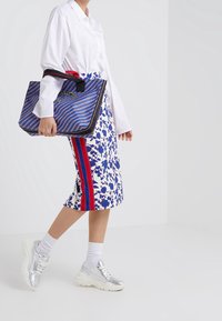 Modrá a bílá květinová tužková sukně se červenými bočními pruhy, spárovaná s bílou košilí na knoflíky a stříbrnými teniskami. Taška má pruhovaný design.