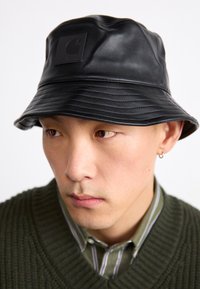 Pălărie din piele neagră, tip găleată, cu un design ribat și un patch pătrat cu logo Carhartt pe față. Dispune de o margine largă, relaxată.