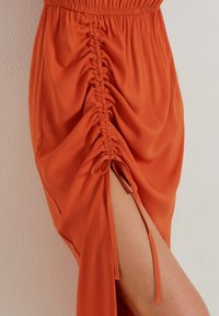 Nařasená oranžová sukně s elastickým pasem a bočním rozparkem. Vyrobená z lehkého materiálu, s texturovanými detaily a vázán�ím.