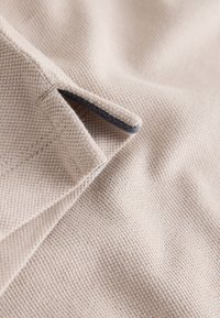 Beige textured fabric with a small stitched loop detail showing a dark underside edge.
