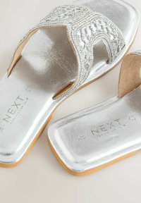 Sandales plates argentées à bouts carrés, brides supérieures ornées de perles et semelles intérieures marron clair, avec le logo "NEXT" sur les semelles.