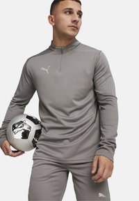 Graues sportliches Oberteil mit einem Reißverschlusskragen und strukturierten Ärmeln, kombiniert mit passenden Hosen. Das Modell hält einen schwarz-weißen Fußball.