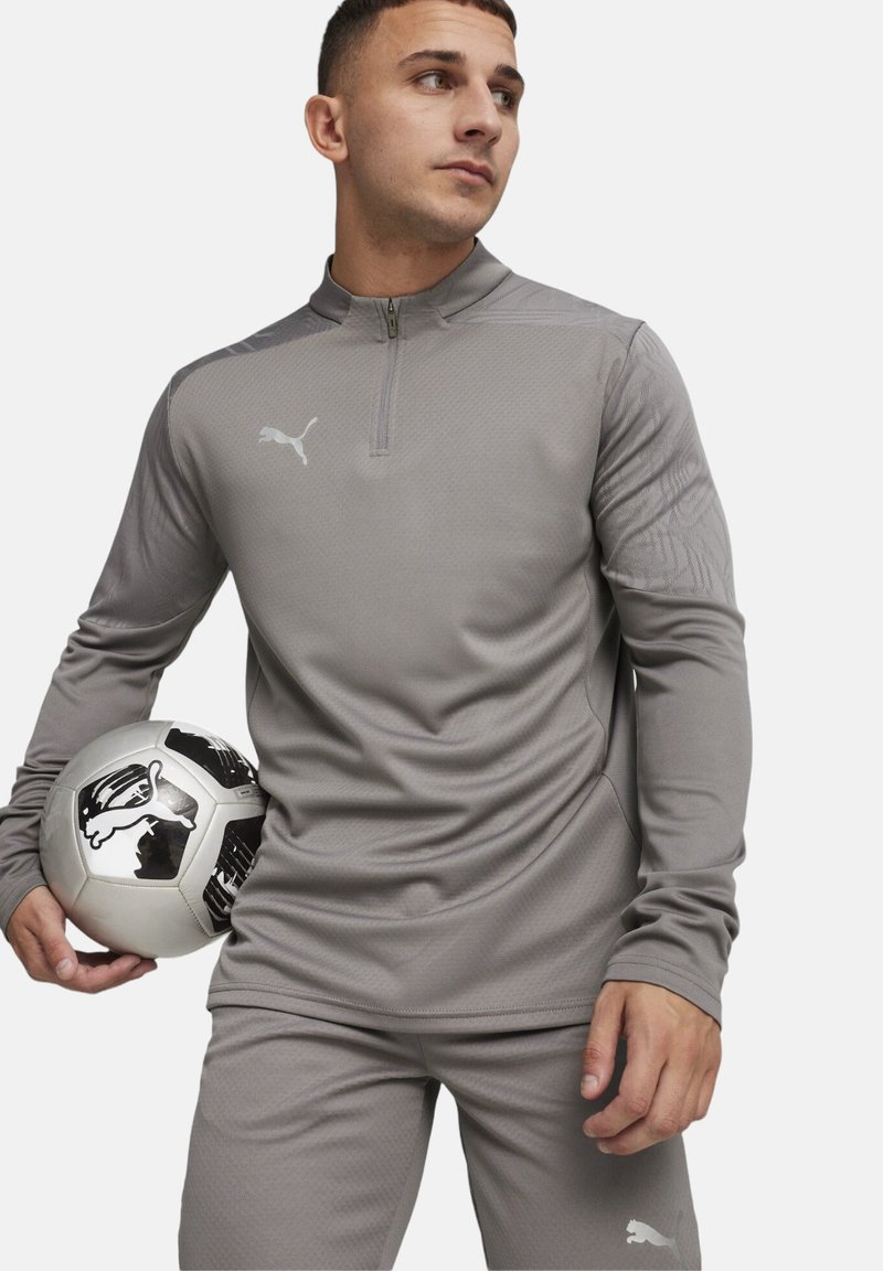 Graues sportliches Oberteil mit einem Reißverschlusskragen und strukturierten Ärmeln, kombiniert mit passenden Hosen. Das Modell hält einen schwarz-weißen Fußball.