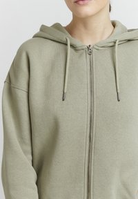 Olivovo zelená mikina na zips z mäkkého, textúrovaného materiálu s kapucňou, strieborným zipsom a šnúrkami, s voľným strihom a spustenými ramennými švami.