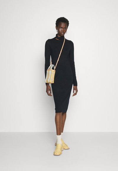 Μαύρο φόρεμα μακρυμάνικο με ψηλό λαιμό και μπροστινό κόψιμο, συνδυασμένο με καρό τσάντα και κίτρινα παπούτσια. Κομψό υλικό, κομψή σιλουέτα.