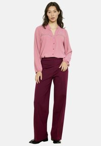 Jasno-różowa bluzka z guzikami, ozdobiona koronką, w zestawie z wysokimi bordowymi spodniami z szerokimi nogawkami i czarnymi butami. Widoczne gładkie faktury materiału.