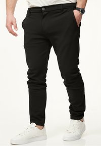 Schwarze Hose aus dehnbarem Material mit slim fit, mit Knopfverschluss, Seitentaschen und aufgerollten Säumen. Kombiniert mit weißen Sneakers.