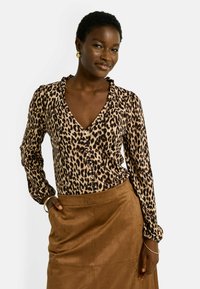Blusa de estampado de leopardo en tejido ligero, con escote en V, cuello con volantes, botón delantero y mangas largas. Combinada con una falda de ante marrón.