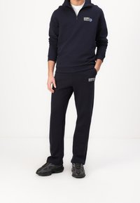 Sudadera marina con media cremallera y logo, combinada con pantalones de chándal marinos a juego que también presentan el logo. La tela es suave; los zapatos son oscuros y texturizados.