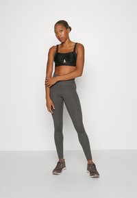 Reggiseno sportivo nero con spalline regolabili e logo, abbinato a leggings grigi aderenti e a vita alta. Il modello indossa scarpe da ginnastica scure.