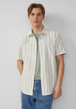 Chemise à manches courtes à boutons de couleur beige clair avec des rayures verticales et une coupe décontractée, superposée à un t-shirt vert pâle, associée à un jean bleu.