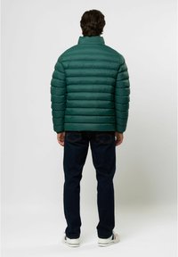 Groene pufferjack met horizontale quilting, hoge kraag en een soepele textuur. Gepaard met donkere jeans en witte schoenen.