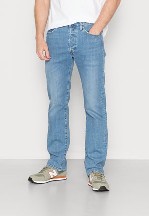 Mann trägt hellblaue Jeans in gerader Passform, Hände in den Taschen, und grau-grüne New Balance Sneakers mit orangefarbenen Akzenten.