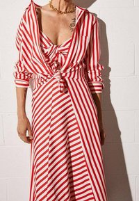 Robe rayée rouge et blanche avec un détail noué à l'avant, manches longues retroussées et une silhouette fluide, fabriquée en tissu léger.