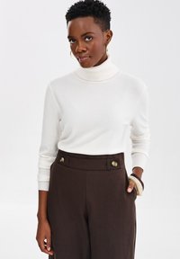 Kobieta z krótkimi włosami, ubrana w biały sweter z golfem i brązowe spodnie z wysokim stanem, z guzikami, uzupełniona bransoletkami i kolczykami w kształcie kół.