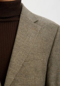 Brun blazer med hundetanns mønster og teksturert stoff, med hakkede slag og brystlomme, brukt over en brun ribbestrikket rullekragegenser.