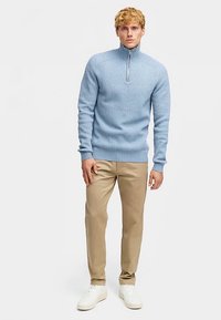 Hellblauer Strickpullover mit Viertel-Reißverschluss am Kragen, gerippter Struktur und figurbetontem Design, kombiniert mit beigen Hosen und weißen Turnschuhen.