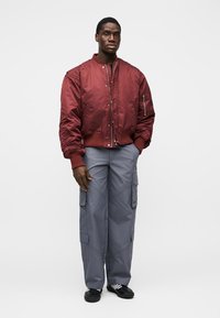 Veste bomber rouge avec poignets côtelés, associée à un pantalon cargo gris avec plusieurs poches. Le modèle se tient les bras détendus le long du corps.