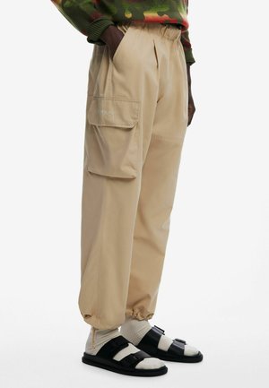 Beige cargo pants with an elastic waistband, pleats, and a side pocket. Paired with black sandals and white socks.
